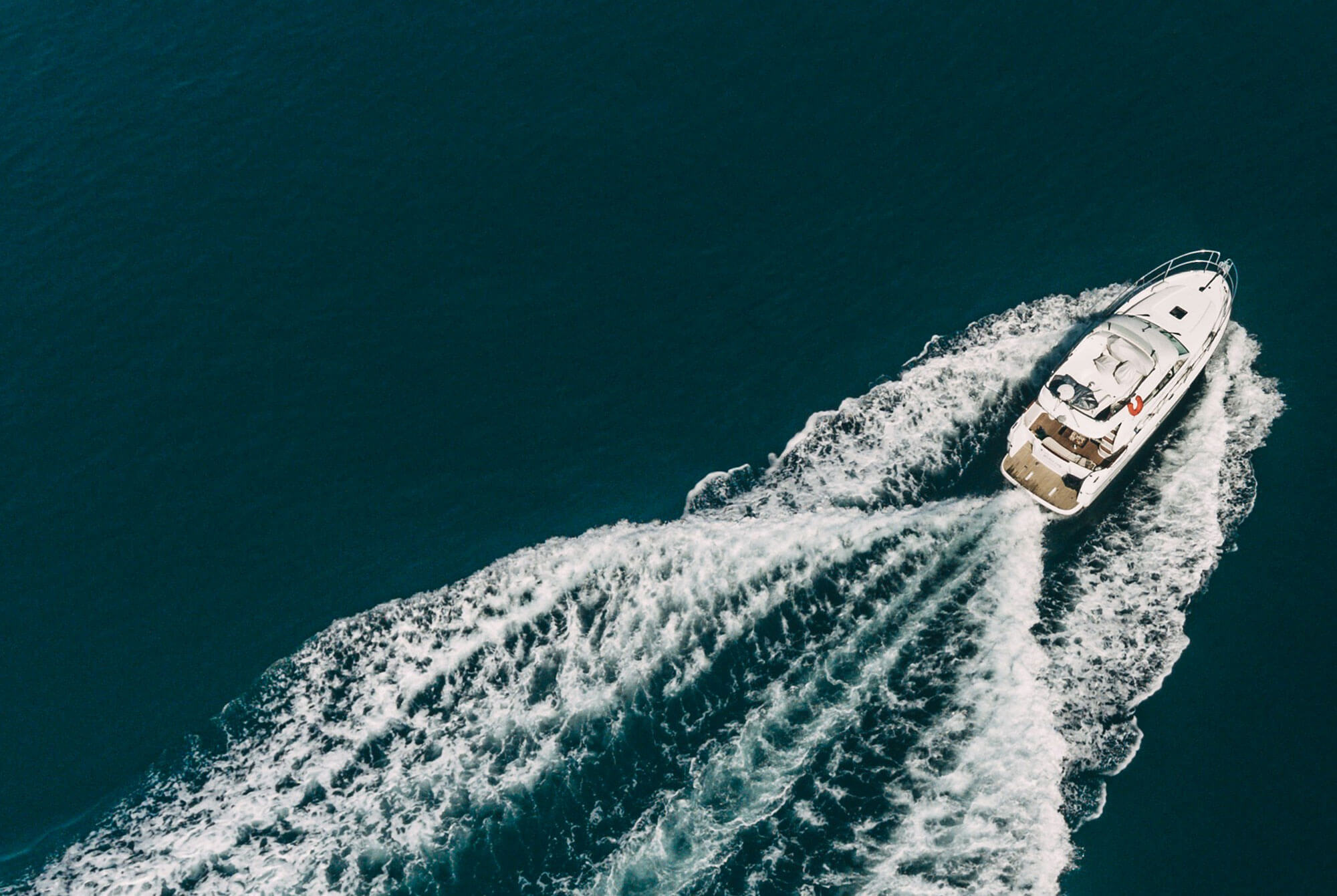 This is an aerial view of a white catamaran boat sailing on dark teal water. The boat is positioned in the bottom-right corner of the image, with people lounging onboard under a small blue sail. The texture and depth of the water dominate the left side, creating a strong contrast between the boat and the vast ocean.